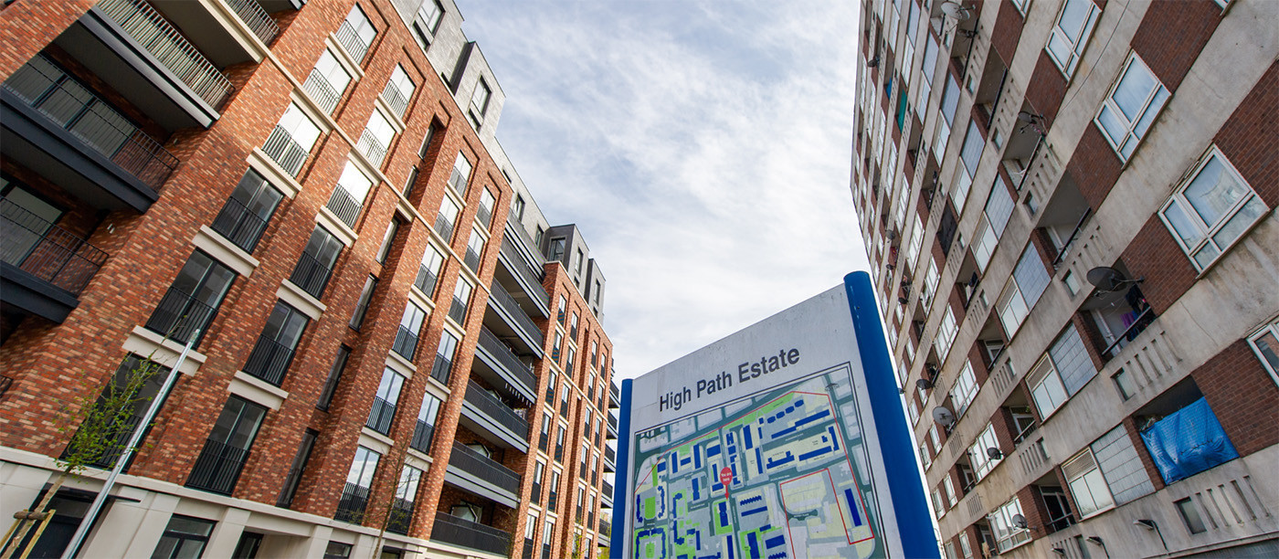 a low-angle photo showing two tower blocks opposite each other, one red brick new-build and one in poor condition. A sign is between the two reading ‘High Path Estate’.